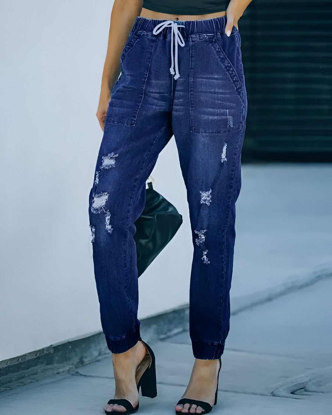 Une femme porte un jean bleu déchiré avec un cordon de serrage à la taille et des talons noirs.