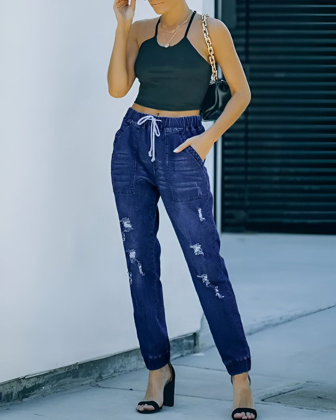 Une jeune femme porte un jean bleu déchiré et un haut noir. Elle est debout dans la rue et a une main dans la poche.