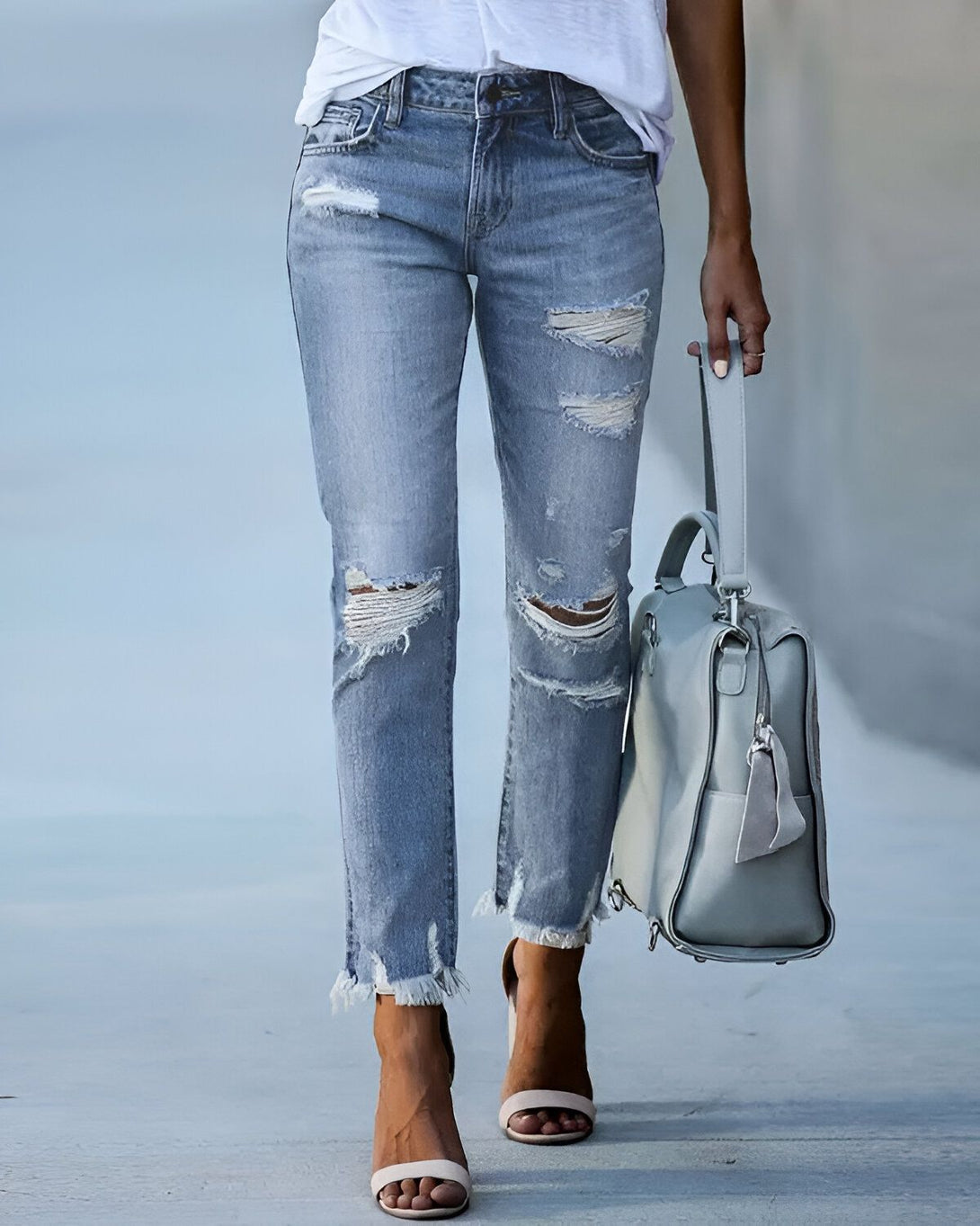 Une femme porte un jean bleu déchiré et troué avec un t-shirt blanc et des sandales à talons hauts. Elle porte un sac à main gris à la main.