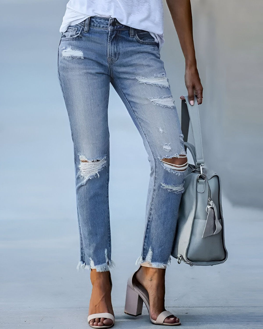 Une femme porte un jean bleu déchiré et troué avec un t-shirt blanc et des sandales à talons hauts. Elle porte un sac à main gris à la main.