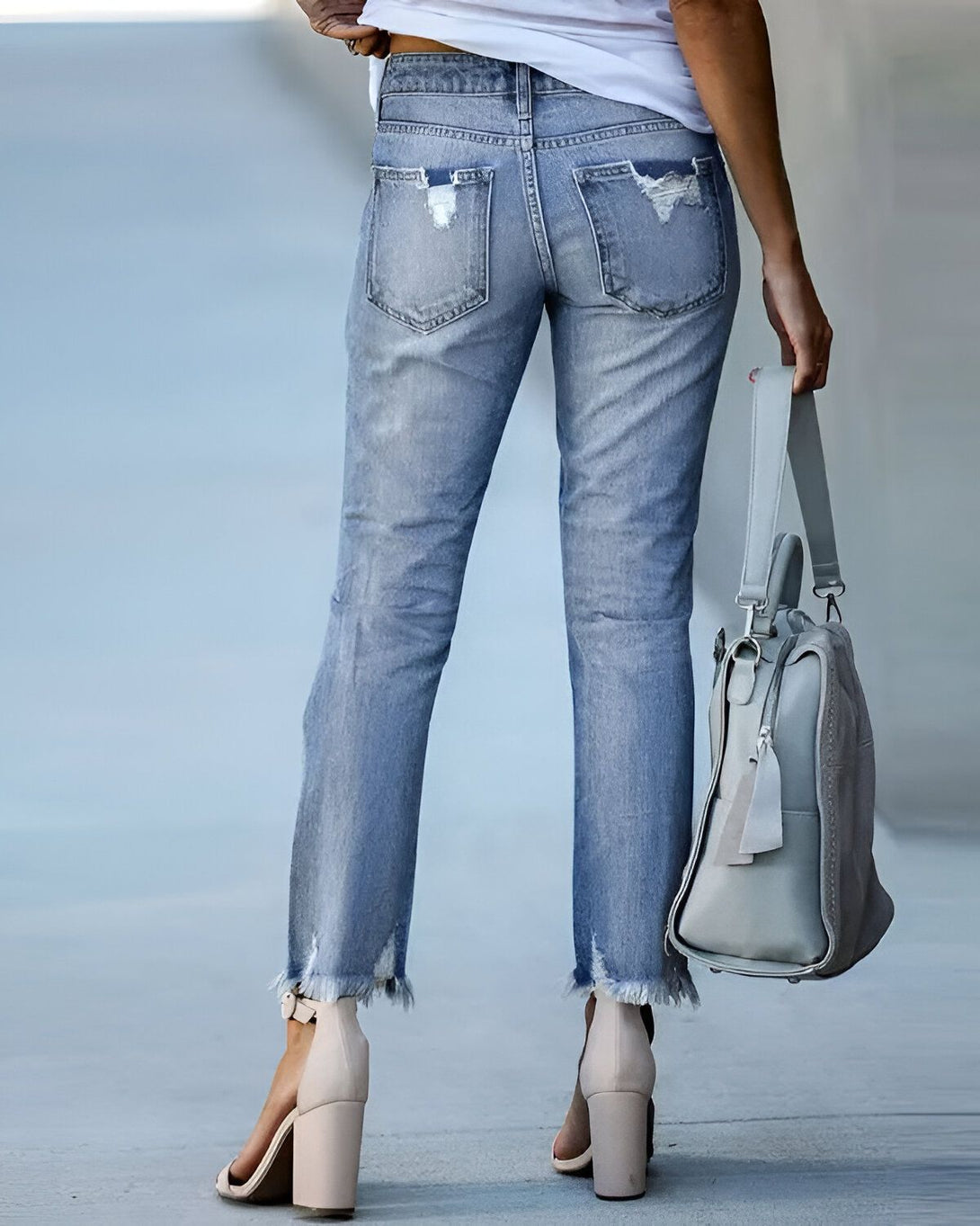 Une femme porte un jean bleu déchiré et des sandales à talons hauts beiges. Elle porte également un sac à main gris.