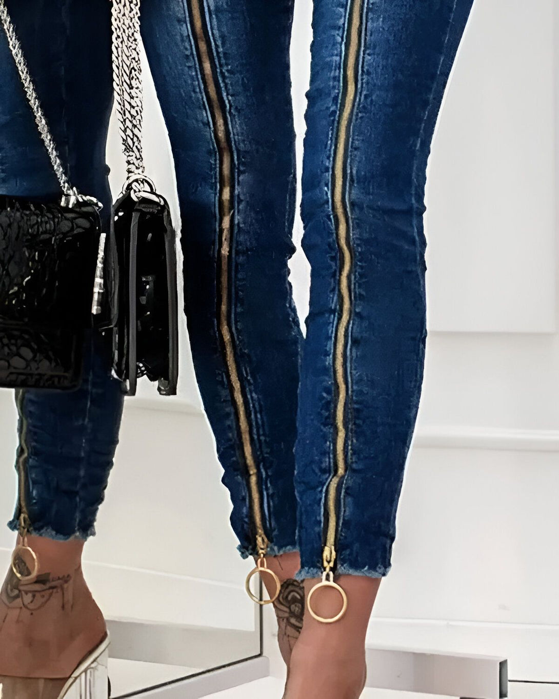 Une femme porte un jean bleu avec des fermetures éclair dorées sur les côtés. Elle porte également des talons hauts transparents et un sac à main noir.