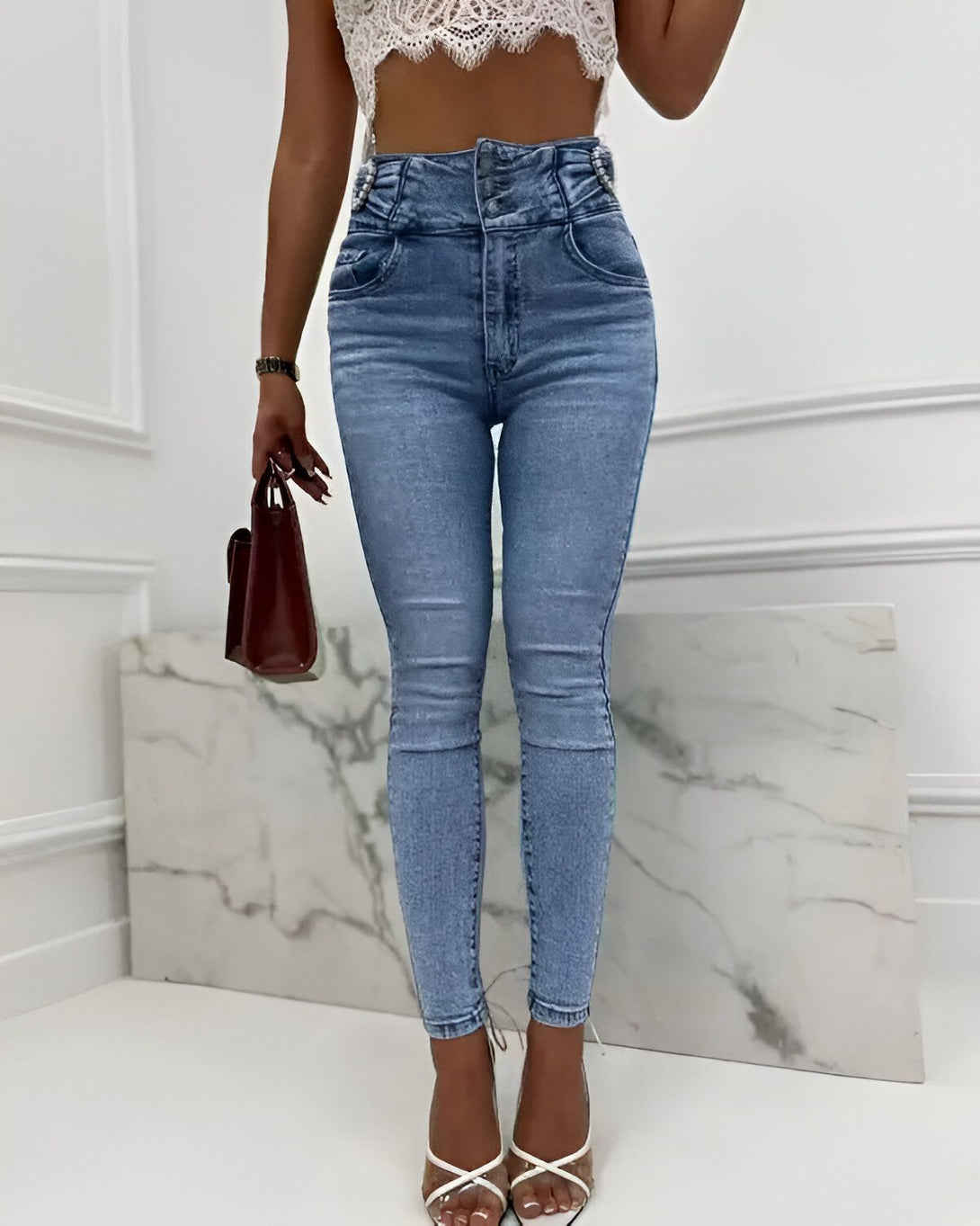 Une femme porte un jean taille haute bleu clair avec des boutons argentés. Elle porte également un haut court en dentelle blanche et des sandales à talons transparentes. Elle a un sac à main marron à la main.