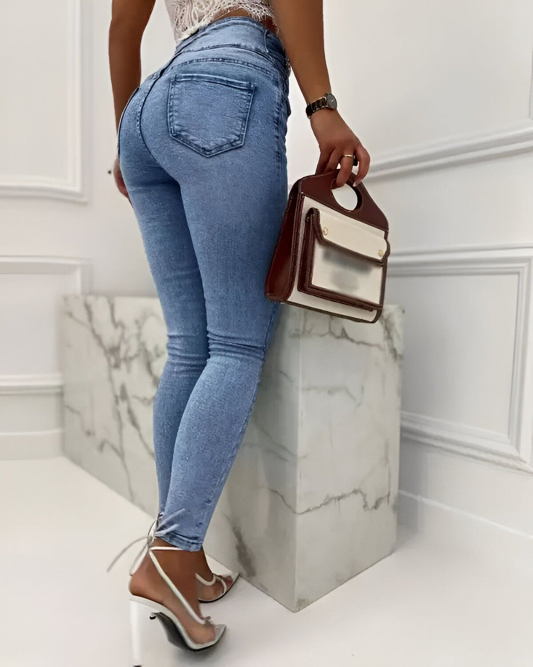 Une femme porte un jean moulant bleu clair et des talons hauts transparents. Elle porte un sac à main marron et blanc à la main droite.