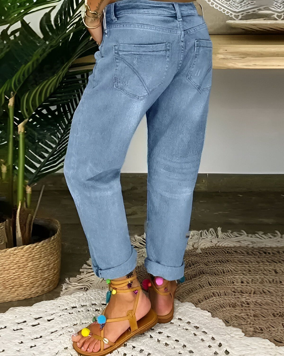 Une femme porte un jean bleu clair et des sandales marron clair. Le jean est retroussé au niveau des chevilles et les sandales sont ornées de pompons colorés. La femme est debout sur un tapis beige et il y a une plante verte à côté d'elle.