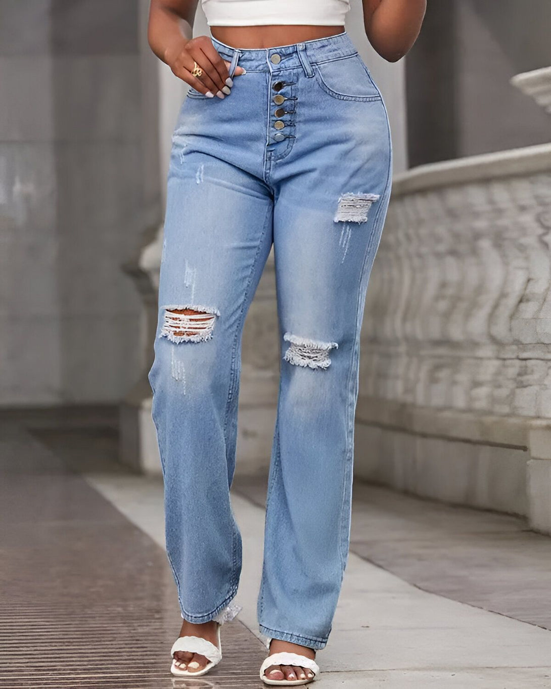 Une femme porte un jean bleu taille haute avec des boutons sur le devant. Le jean est déchiré au niveau des genoux. Elle porte des sandales blanches à talons.