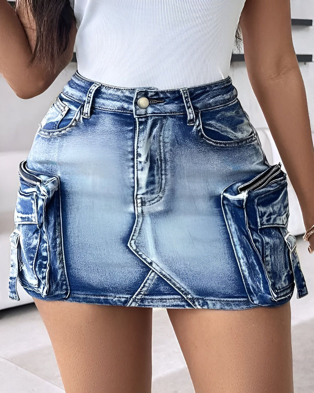 Une jeune femme porte une minijupe en jean bleu clair. La jupe est taille haute et possède des poches zippées sur les côtés. La femme porte un débardeur blanc et des talons hauts noirs.