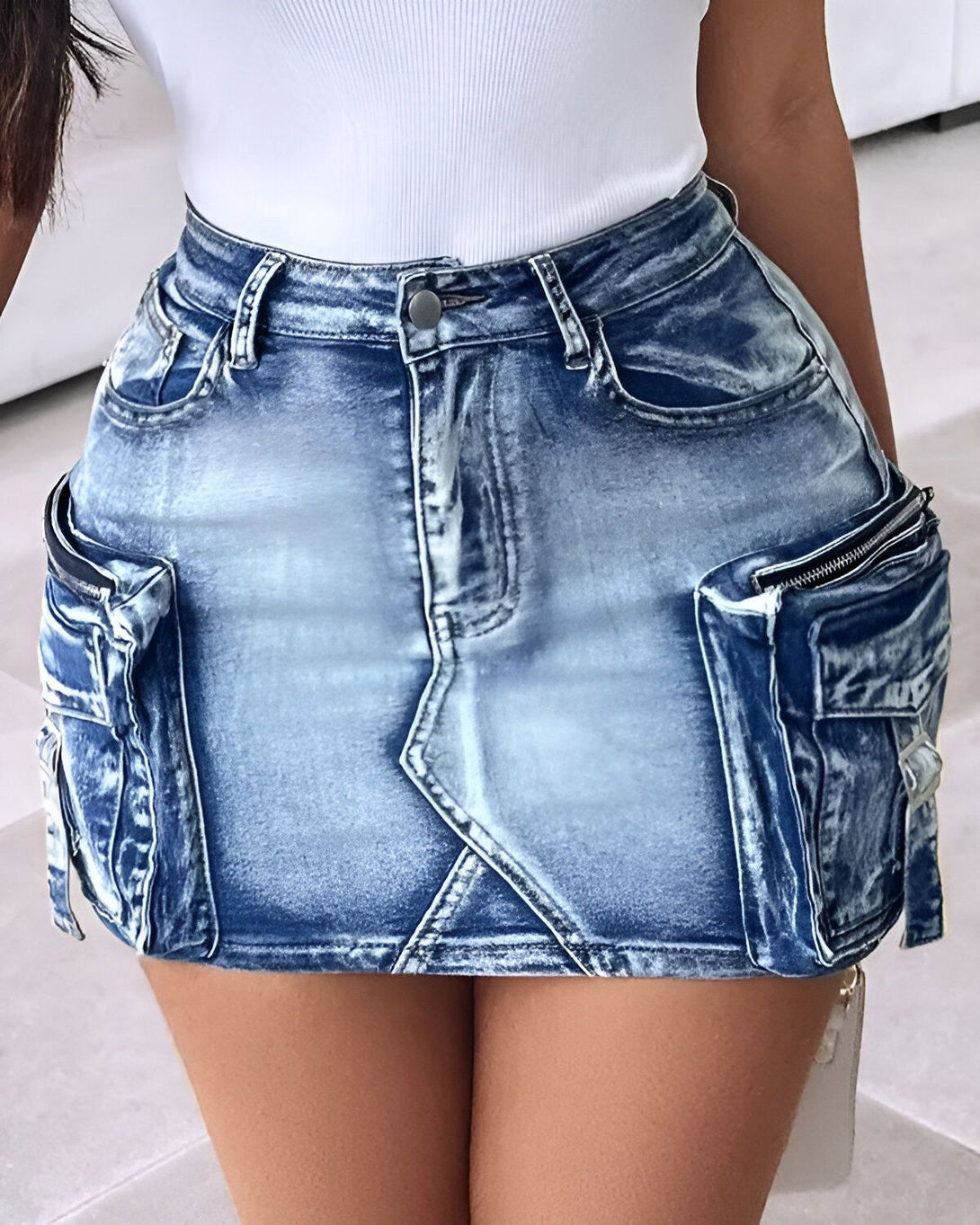 Une jeune femme porte une minijupe en jean bleu clair. La jupe est taille haute et possède des poches cargo sur les côtés. La femme porte un débardeur blanc et des talons hauts noirs.
