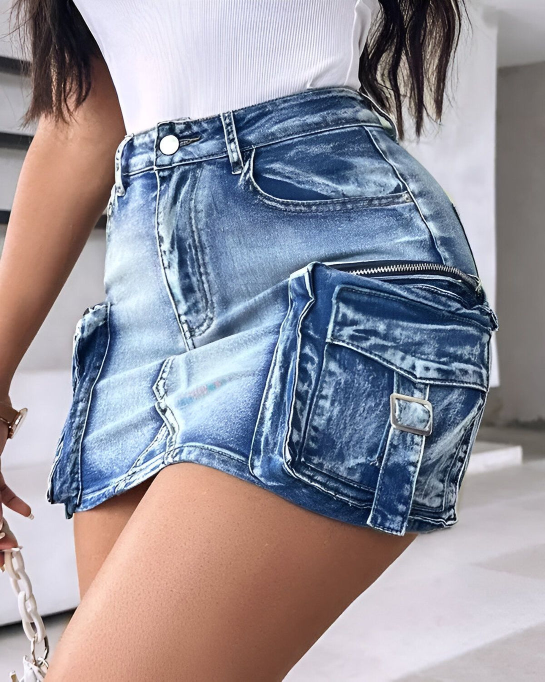 Une jeune femme porte une mini-jupe en jean bleu clair. La jupe est taille haute et possède des poches cargo sur les côtés. La femme est vue de dos et porte un haut blanc.