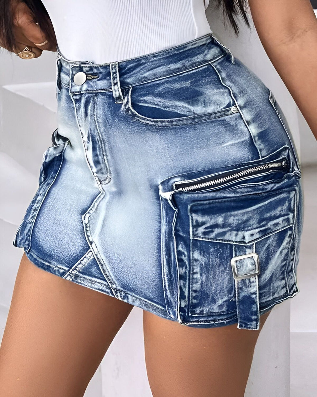Une jeune femme porte une mini-jupe en jean bleu clair. La jupe est taille haute et possède des poches cargo sur les côtés. Elle est associée à un débardeur blanc.