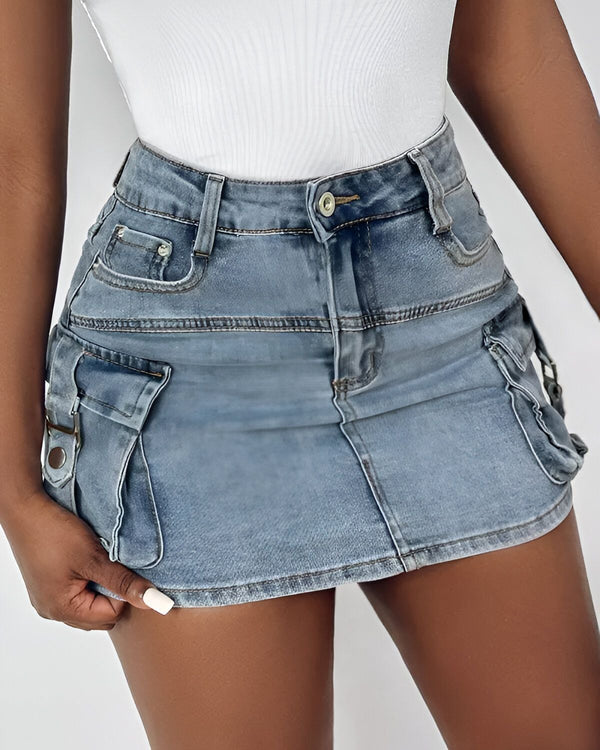 Une jeune femme porte une minijupe en jean bleu clair. La jupe est taille haute et possède des poches cargo sur les côtés. La femme porte également un débardeur blanc.
