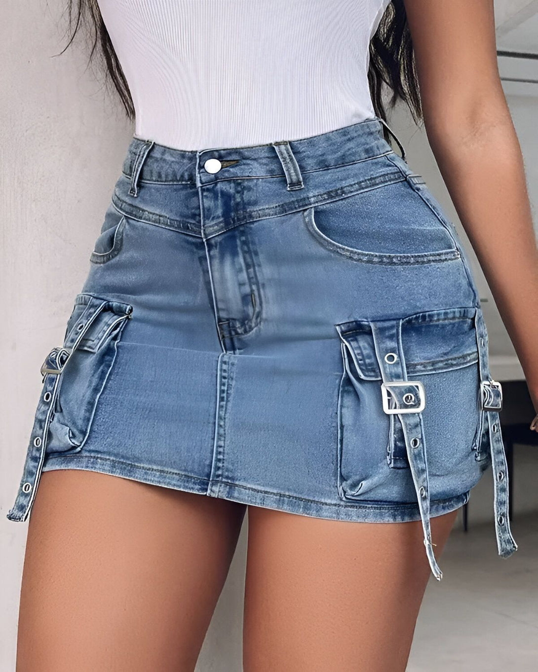 Une jeune femme porte une mini-jupe en jean bleu clair. La jupe est taille haute et possède des poches cargo sur les côtés. Elle est associée à un débardeur blanc.