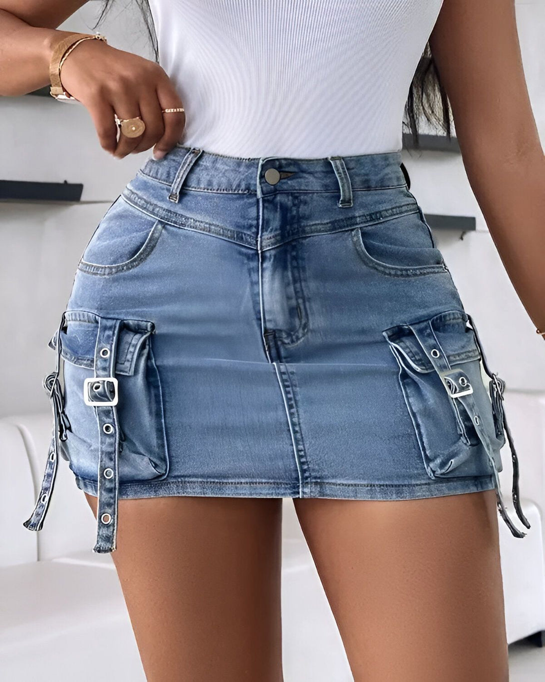 Une jeune femme porte une mini-jupe en jean bleu clair. La jupe est taille haute et possède des poches cargo sur les côtés. Elle est associée à un débardeur blanc.