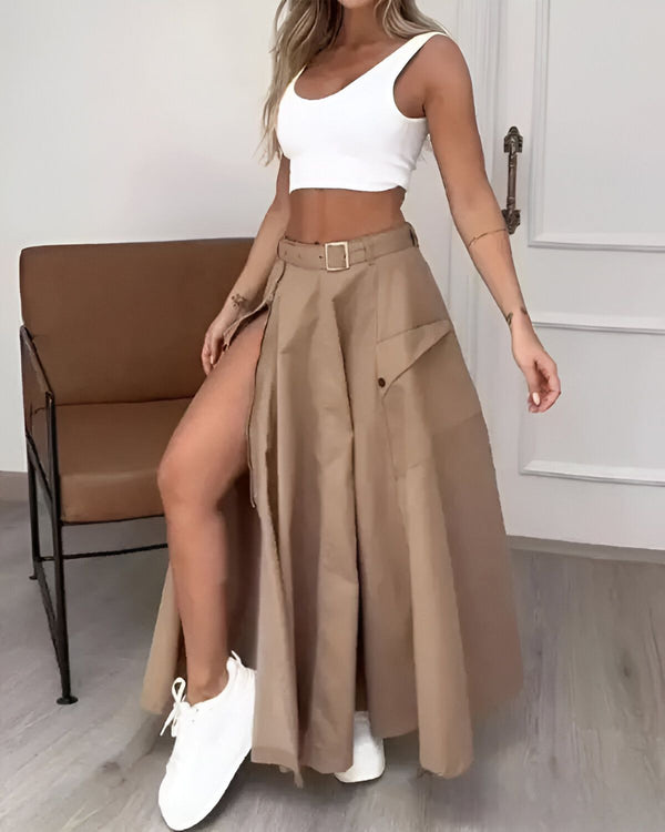 Une femme porte une jupe longue marron fendue sur le côté, un crop top blanc et des baskets blanches.
