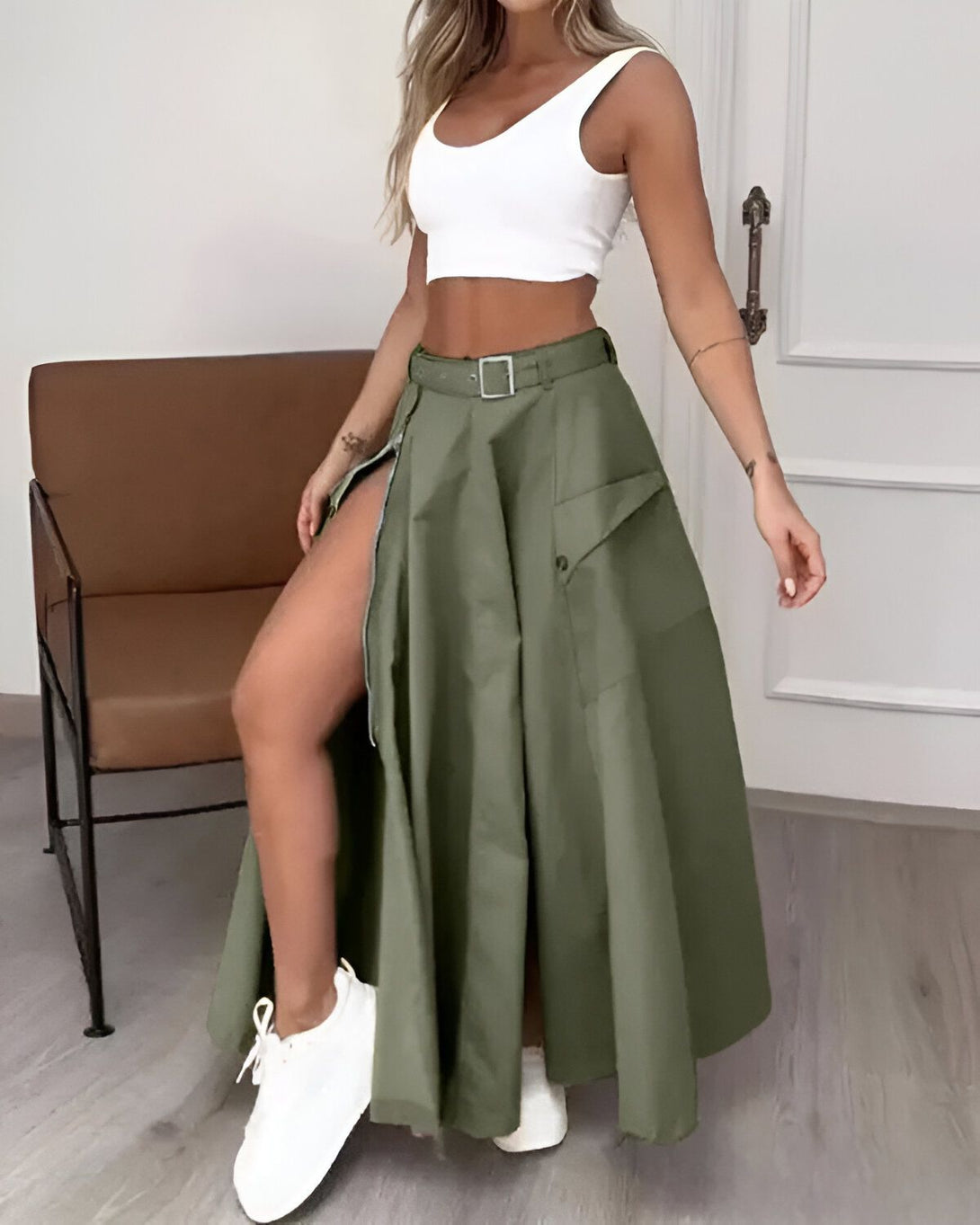 Une femme porte une jupe longue kaki fendue sur le côté avec une ceinture à la taille. Elle porte également un débardeur blanc et des baskets blanches.