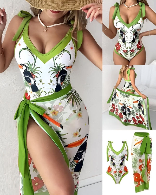 Une femme porte un maillot de bain une pièce avec un imprimé tropical. Le maillot de bain est vert et blanc, avec des fleurs et des oiseaux. La femme porte également un chapeau de paille et un paréo assorti.