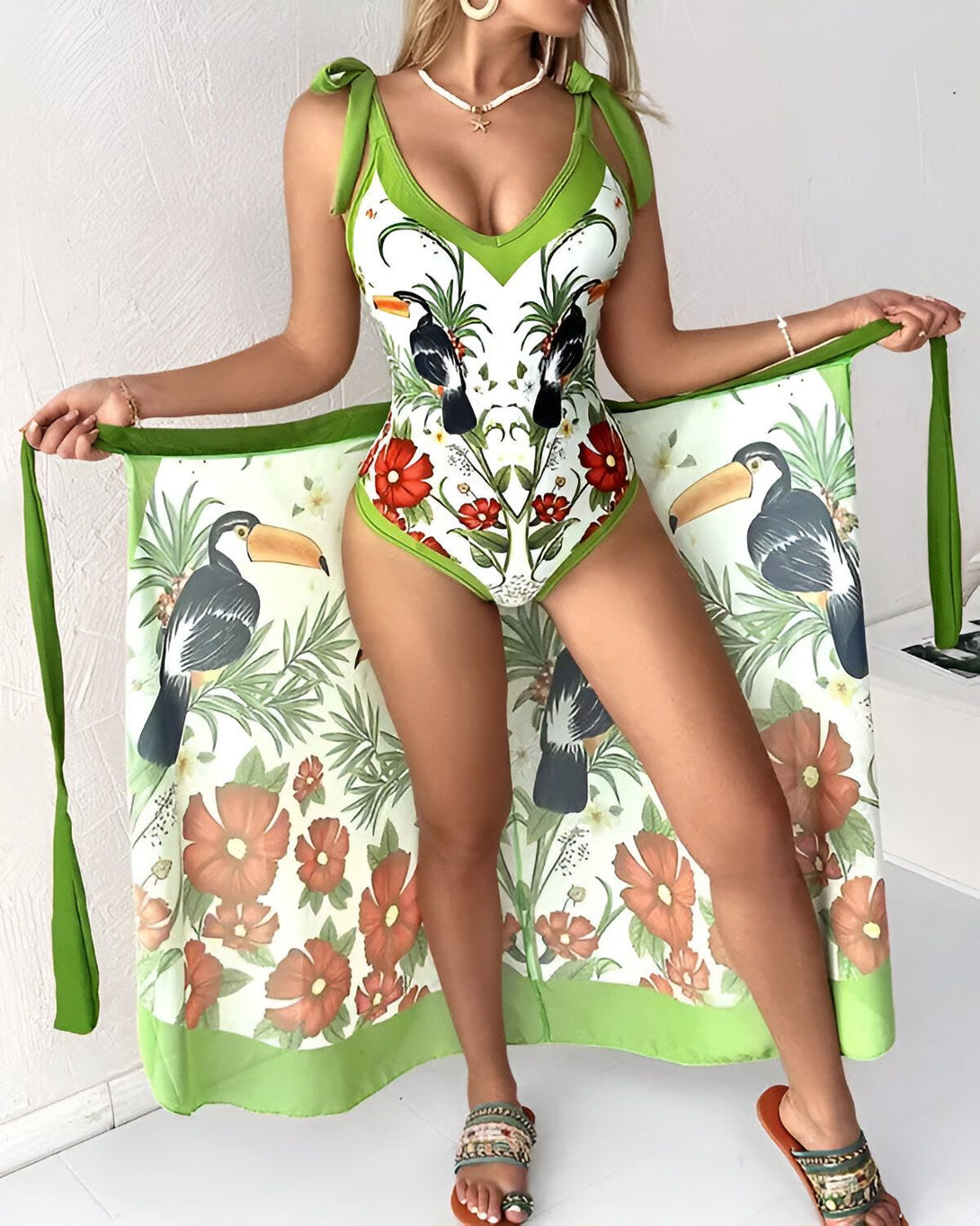 Une femme porte un maillot de bain une pièce vert avec un motif de fleurs et de toucans. Elle porte également une paréo assorti.