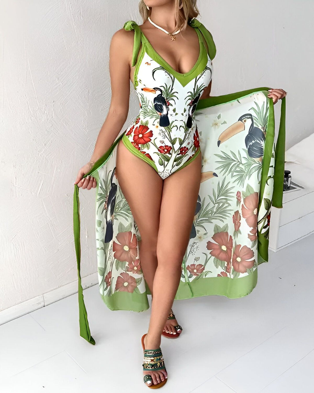 Une femme porte un maillot de bain une pièce vert avec un imprimé de fleurs et de toucans. Elle porte également une longue robe de plage verte assortie.