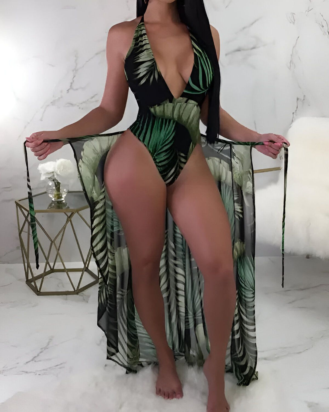 Une femme portant un maillot de bain une pièce noir et vert à motifs de feuilles de palmier. Elle porte également une longue robe de plage transparente à motifs de feuilles de palmier.