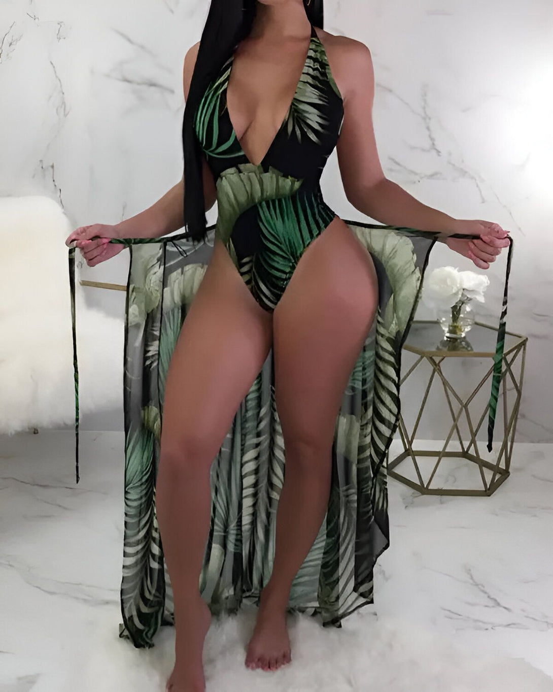 Une femme porte un maillot de bain une pièce noir avec un imprimé de feuilles tropicales vertes. Elle porte également une longue robe de plage transparente à imprimé de feuilles tropicales vertes.