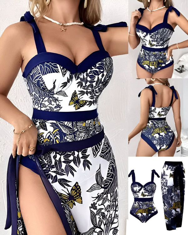 Une femme porte un maillot de bain une pièce bleu marine et blanc à motifs de papillons et de fleurs. Le maillot de bain est échancré sur les côtés et possède un lien à nouer autour du cou. La femme porte également une paréo assorti.