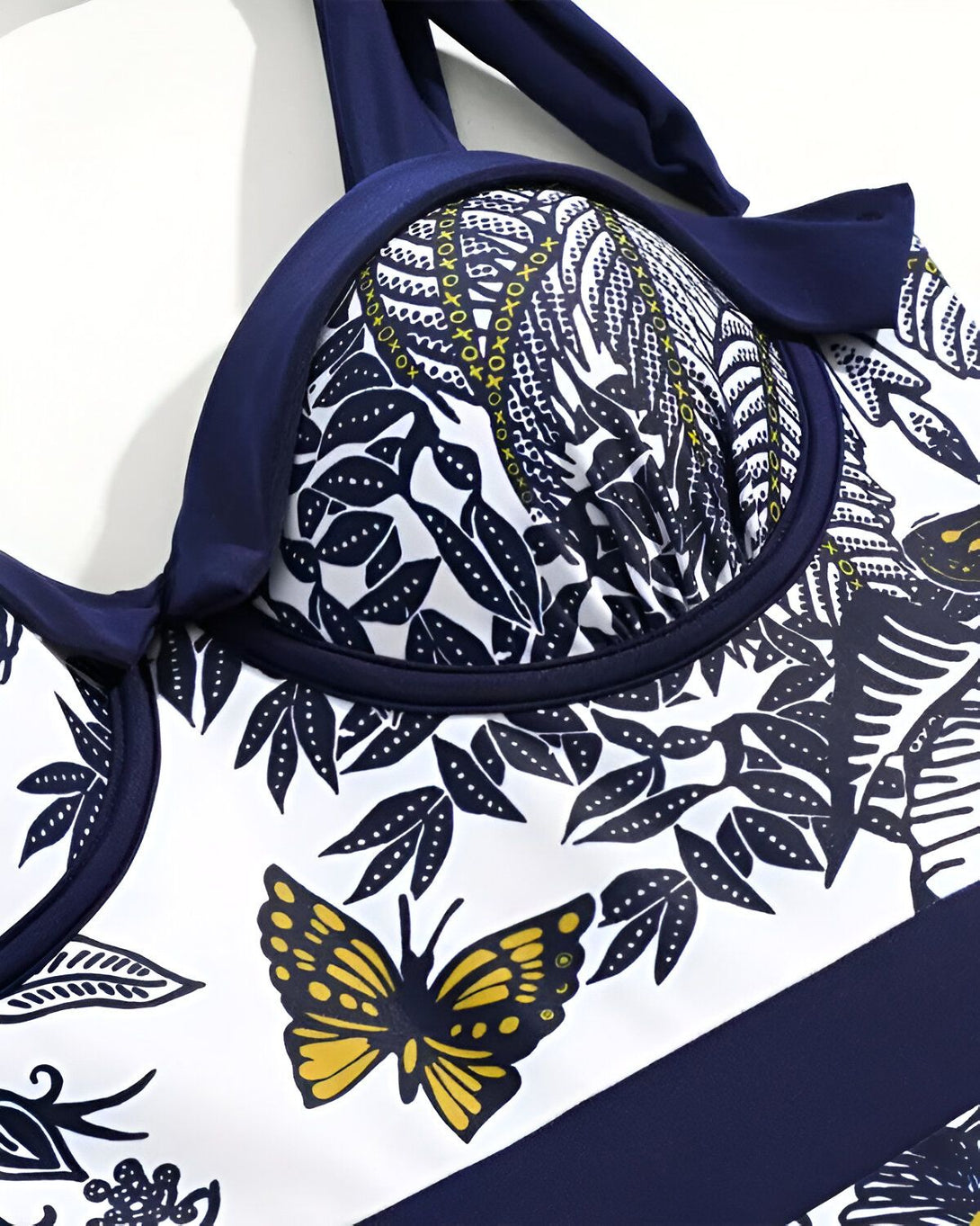 Un maillot de bain une pièce avec un imprimé de feuilles tropicales et de papillons. Le haut du maillot de bain est bleu marine avec des bonnets rembourrés et des bretelles fines. Le bas du maillot de bain est blanc avec une taille haute et des échancrures modérées.