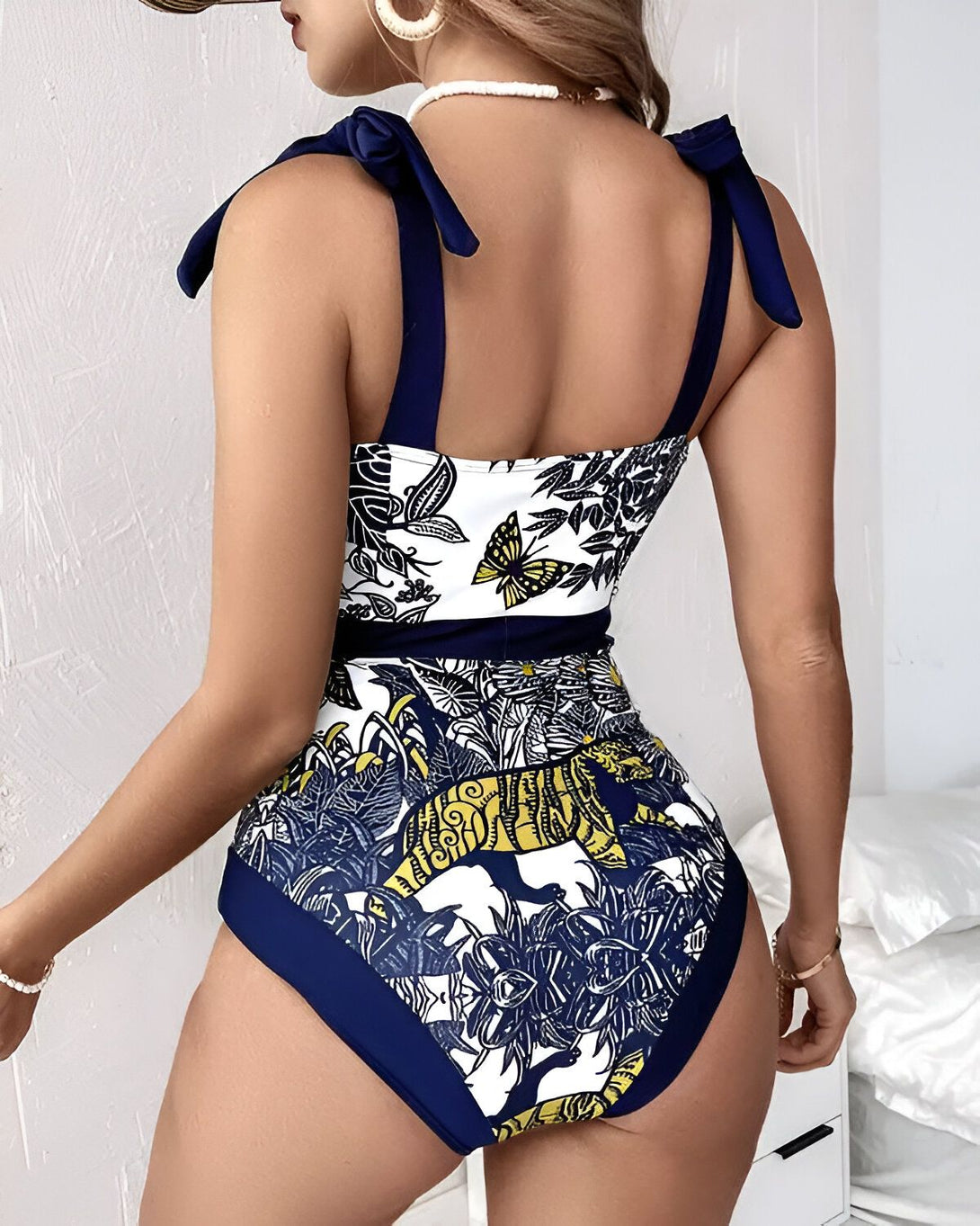 Une femme porte un maillot de bain une pièce bleu et jaune. Le maillot de bain a un motif de feuilles et de fleurs tropicales. Il est échancré sur les côtés et a un dos nageur. La femme est vue de dos.