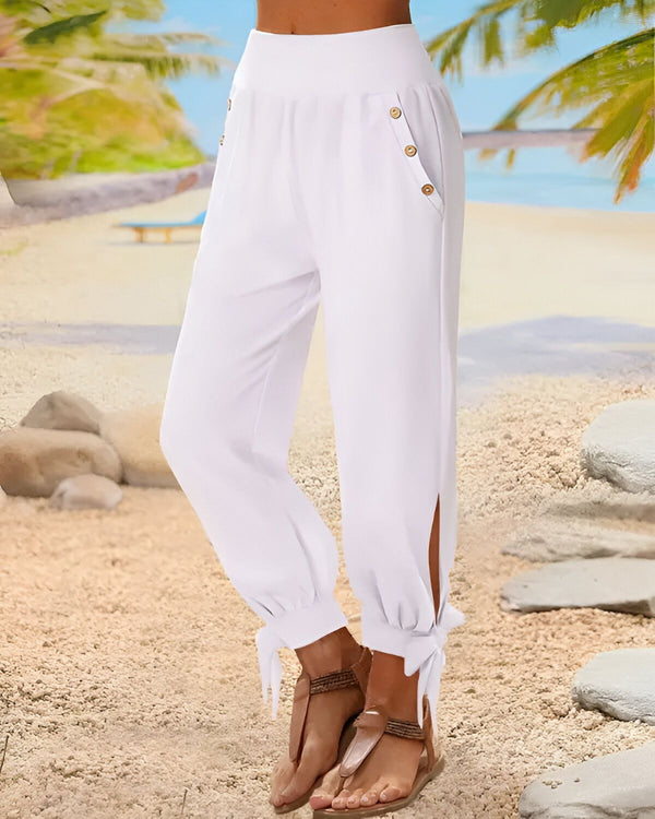 Une femme porte un pantalon blanc ample et fluide, avec des boutons sur le devant et des liens aux chevilles. Elle est pieds nus et se trouve sur une plage, avec des palmiers et la mer en arrière-plan.