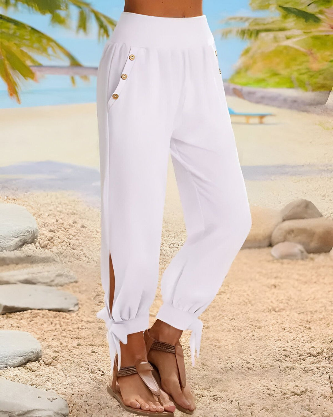 Une femme porte un pantalon blanc ample et léger. Le pantalon est noué au niveau des chevilles et possède des boutons décoratifs sur le devant. La femme est pieds nus et porte des sandales marron clair. Elle se trouve sur une plage de sable blanc avec des palmiers en arrière-plan.
