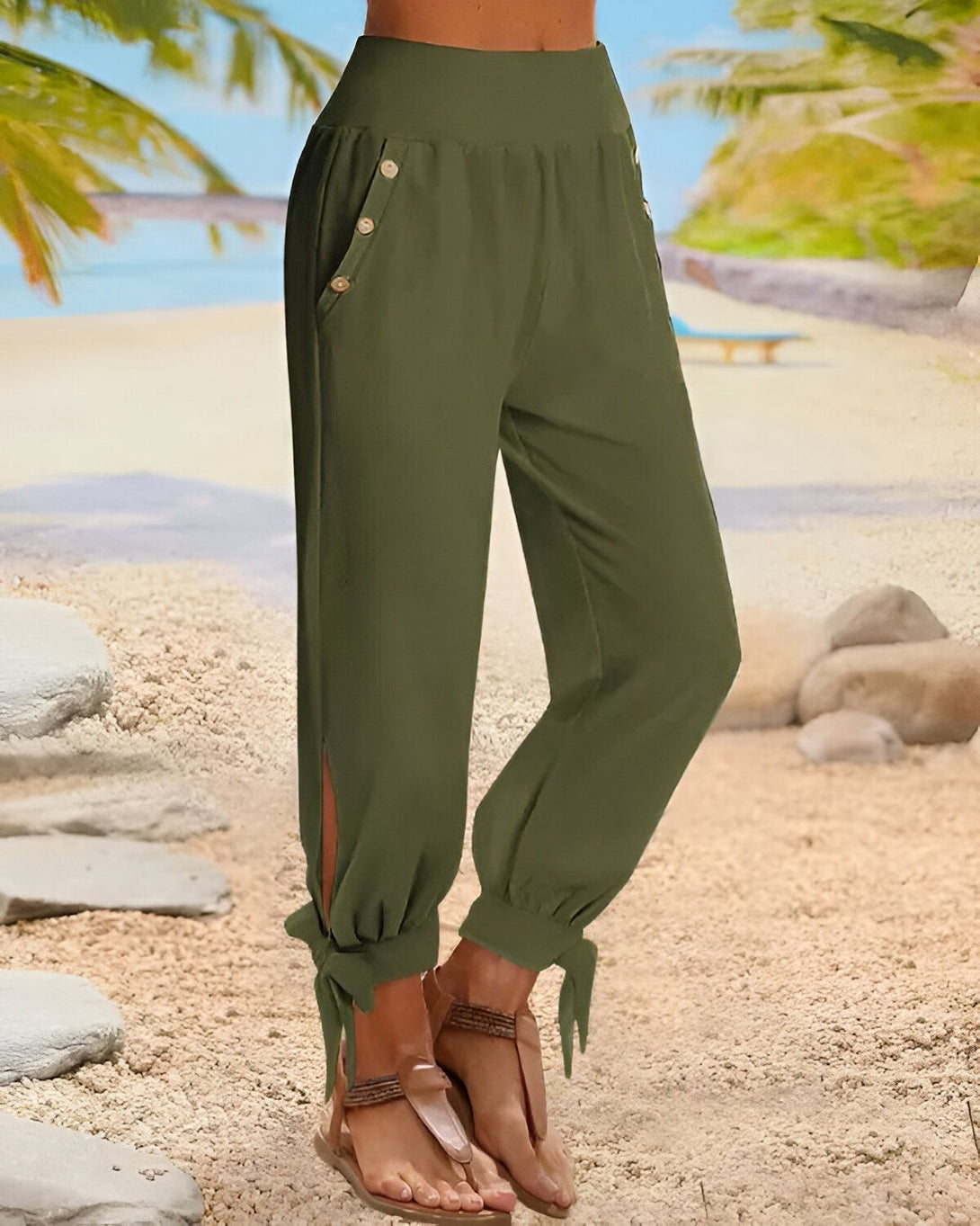 Une femme porte un pantalon vert olive taille haute avec des boutons sur le côté. Le pantalon est ample et a des jambes larges. La femme est pieds nus et porte des sandales marron clair. Elle se tient sur une plage de sable fin avec des palmiers en arrière-plan.