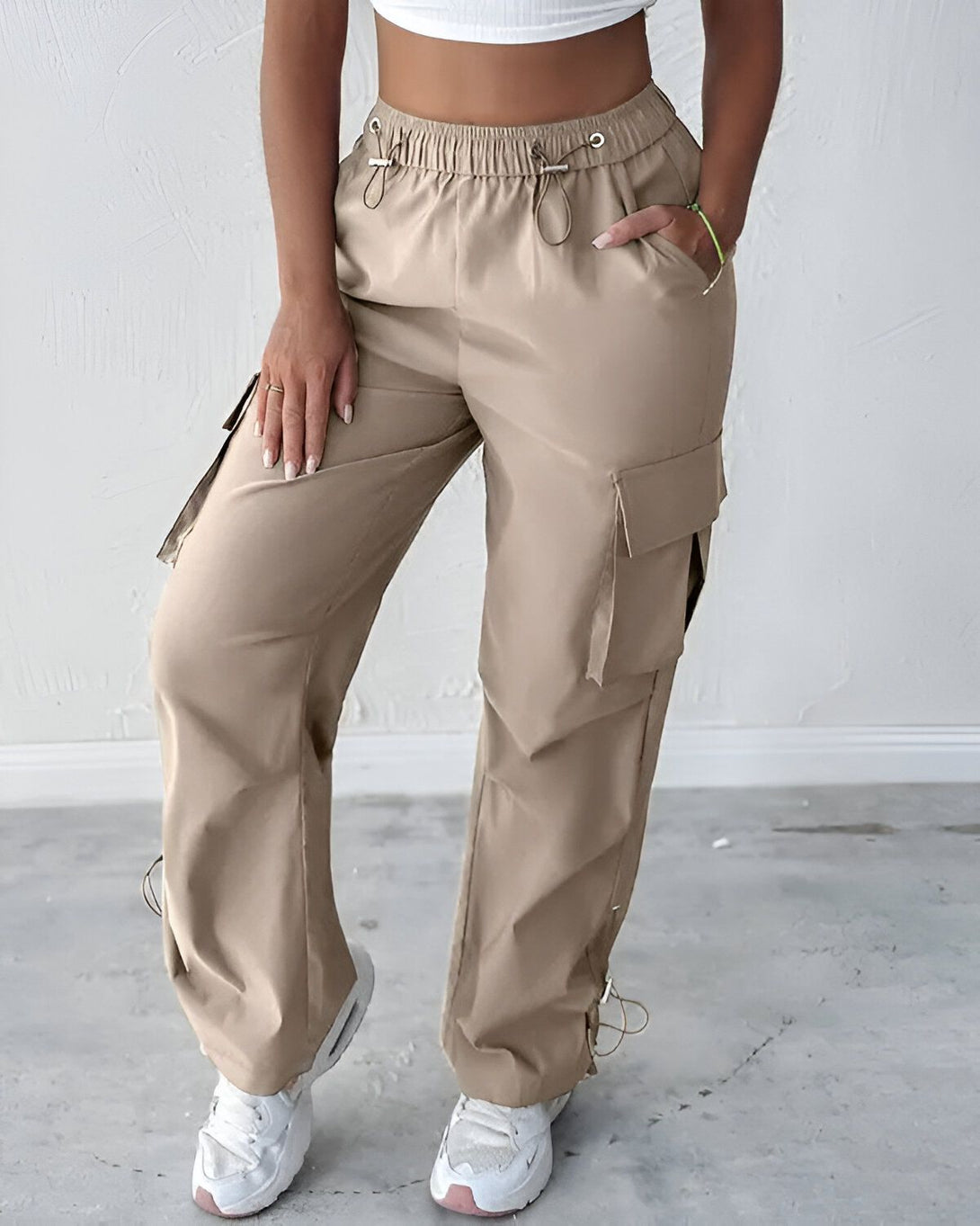 Une femme porte un pantalon cargo beige taille haute avec des poches sur les côtés. Elle porte également un crop top blanc et des baskets blanches.