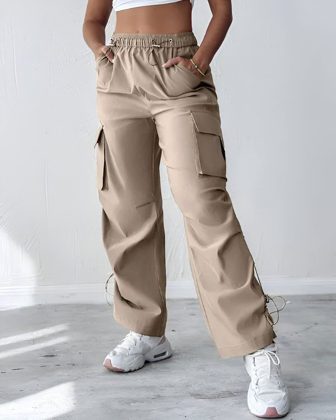 Une femme porte un pantalon cargo beige ample avec des poches cargo sur les côtés. Elle porte également un haut court blanc et des baskets blanches.
