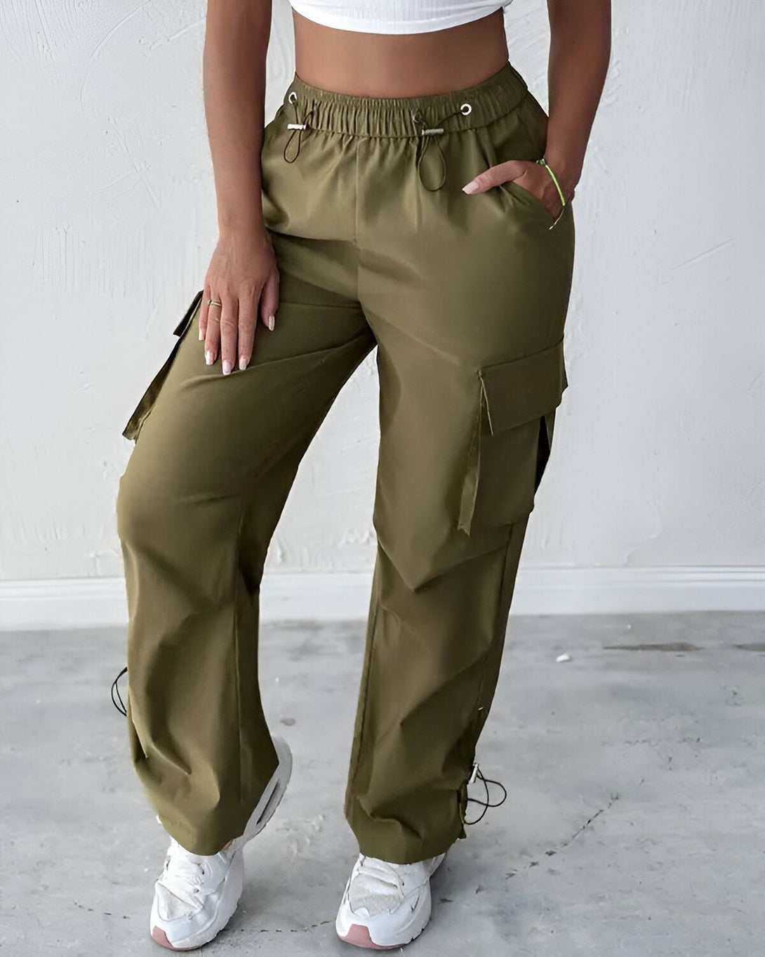 Une femme porte un pantalon cargo vert olive taille haute avec des poches cargo sur les côtés. Elle porte également un crop top blanc et des baskets blanches.