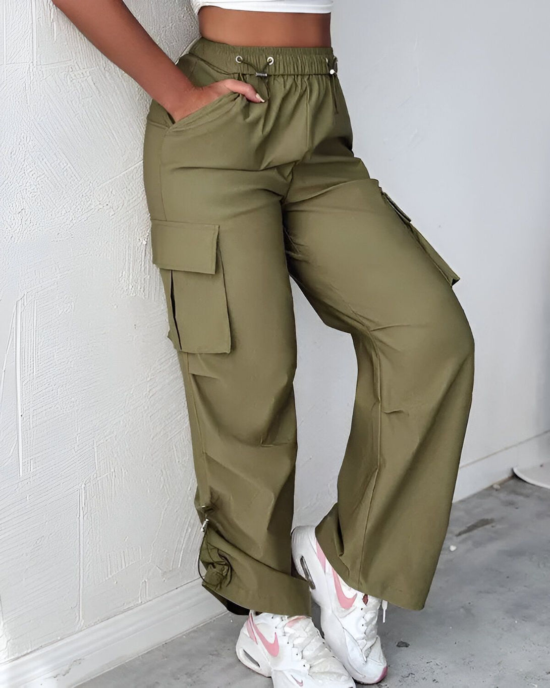 Une femme porte un pantalon cargo vert olive taille haute avec des poches cargo sur les côtés. Elle porte également un haut court blanc et des baskets blanches.