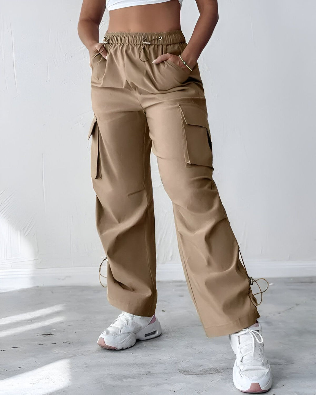 Une femme porte un pantalon cargo beige ample avec des poches et des cordons de serrage à la taille et aux chevilles. Elle porte également un haut court blanc et des baskets blanches.