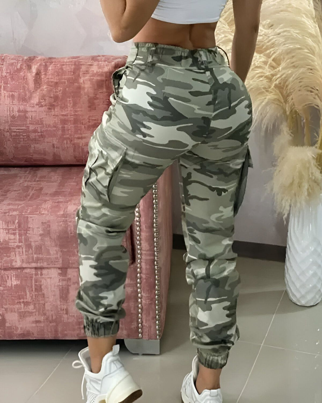 Une femme porte un pantalon cargo camouflage vert et blanc avec une taille haute et des poches cargo sur les côtés. Elle porte également un haut blanc court et des baskets blanches.