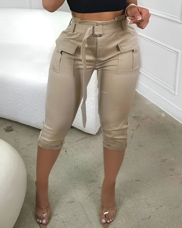 Une femme porte un pantalon cargo beige taille haute. Le pantalon est ceinturé et possède des poches sur les côtés. La femme porte également un haut noir et des sandales transparentes.
