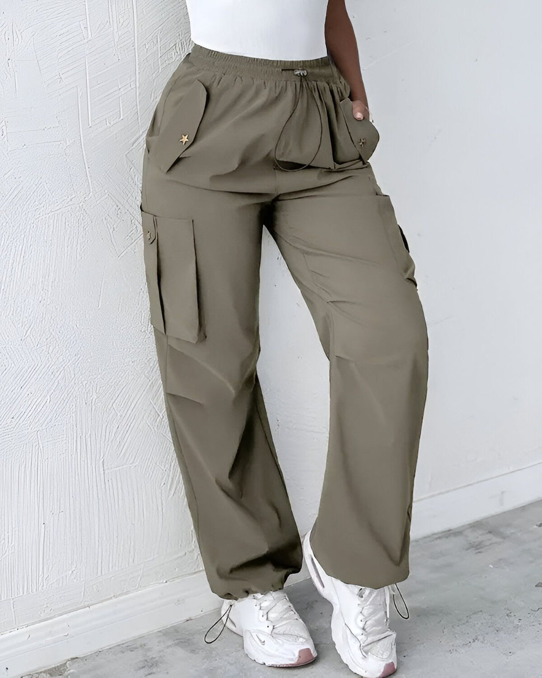 Une femme porte un pantalon cargo vert olive taille haute avec des poches cargo sur les côtés. Elle porte également un haut blanc et des baskets blanches.