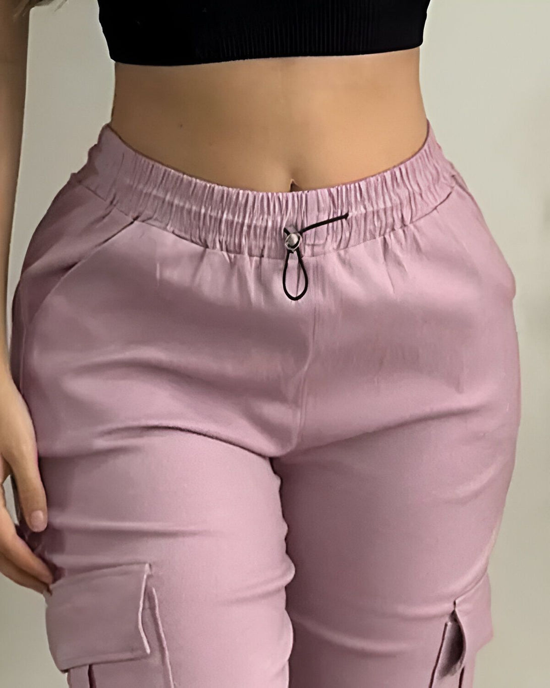 Une femme porte un pantalon cargo rose et un haut noir. Le pantalon est taille haute et possède des poches cargo sur les côtés. La femme est vue de face et elle a les cheveux longs et noirs.