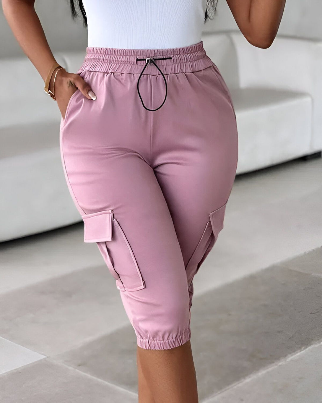 Une femme porte un pantalon cargo rose. Le pantalon est taille haute et possède une coupe ample. Il est doté de deux poches cargo sur les côtés et d'un cordon de serrage à la taille. La femme porte également un débardeur blanc et des sandales noires.