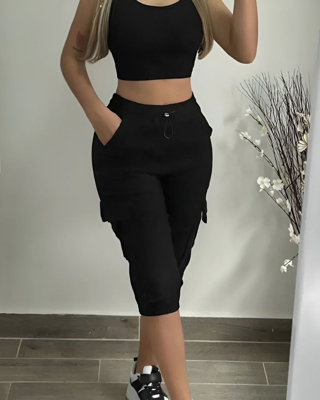 Une femme porte un haut court noir et un pantalon cargo noir. Elle est debout dans un miroir et tient son téléphone dans sa main droite.