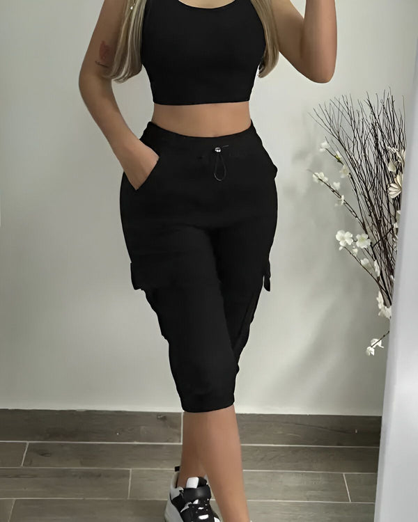 Une femme porte un haut court noir et un pantalon cargo noir. Elle est debout dans un miroir et tient son téléphone dans sa main droite.