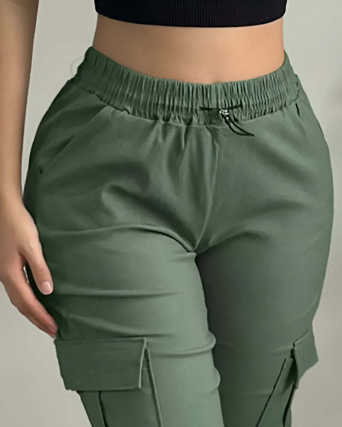 Une femme porte un pantalon cargo vert olive taille haute. Le pantalon est ample et a des poches cargo sur les côtés. La femme porte également un haut noir court.