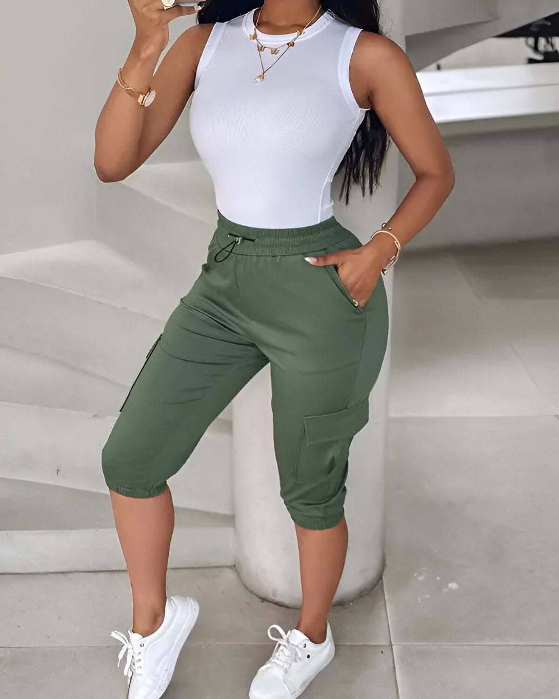 Une femme porte un débardeur blanc et un pantalon cargo vert olive. Elle est debout dans un escalier et tient son téléphone dans sa main droite.