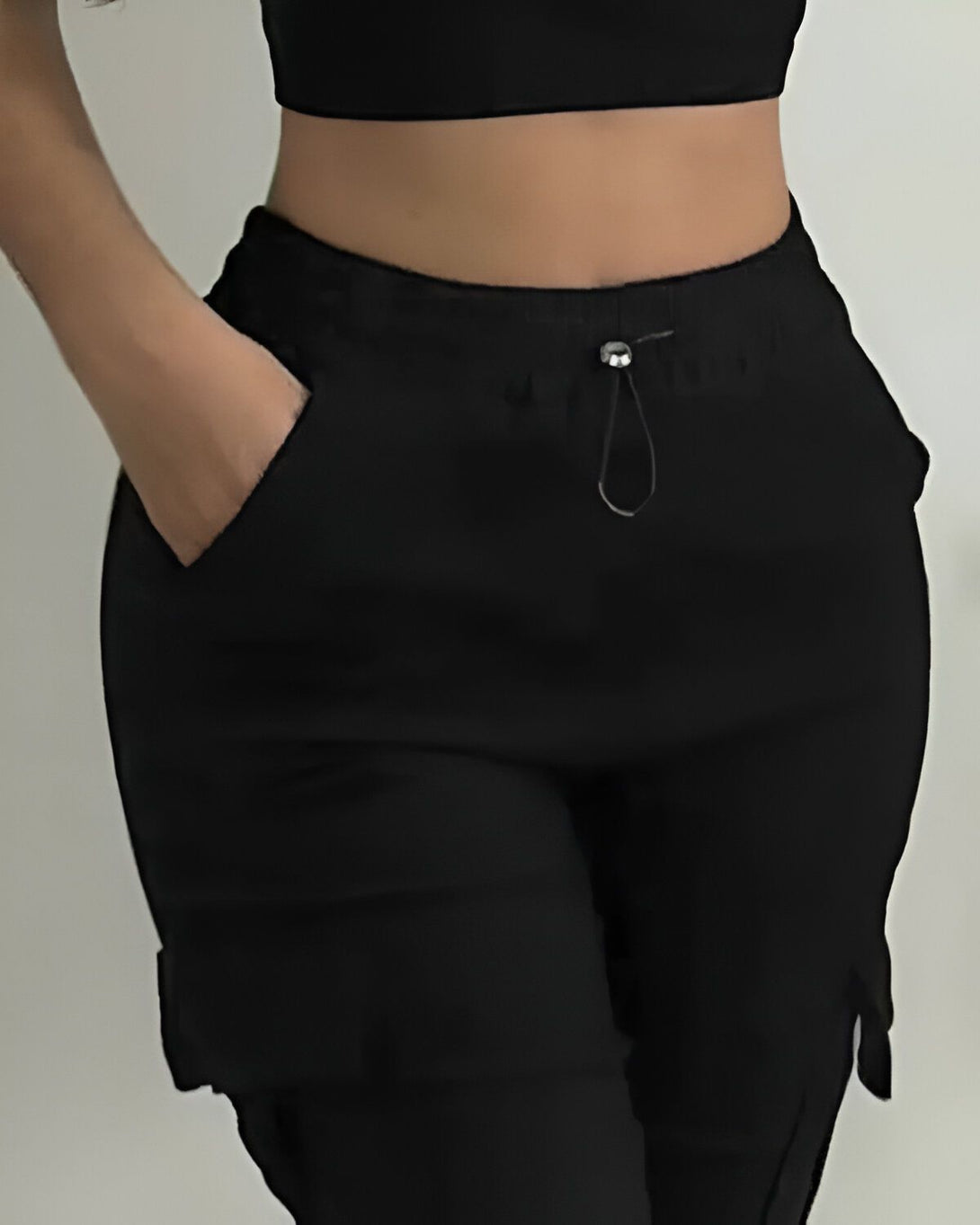 Une femme porte un pantalon cargo noir taille haute avec une poche sur le côté. Elle porte également un haut court noir.