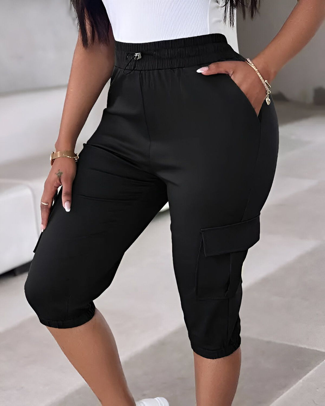 Une femme porte un short cargo noir. Le short est taille haute et a une coupe ample. Il possède des poches cargo sur les côtés et un cordon de serrage à la taille. La femme porte également un débardeur blanc et des baskets blanches.