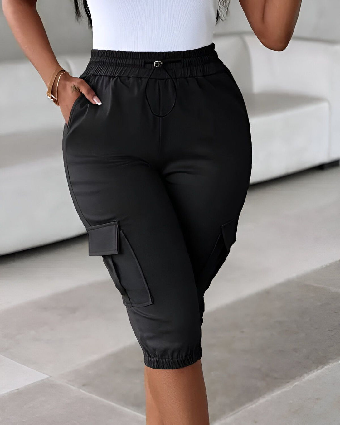 Une femme porte un pantalon cargo noir. Le pantalon est taille haute et a une coupe ample. Il est doté de poches cargo sur les côtés et d'un cordon de serrage à la taille. La femme porte également un débardeur blanc et des talons noirs.