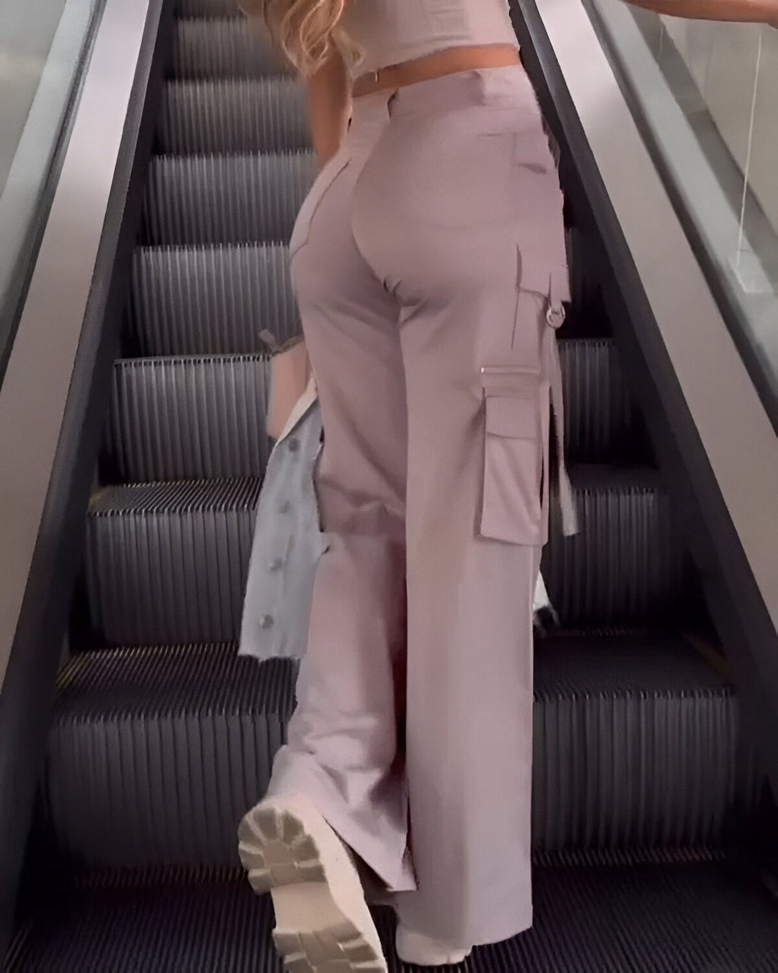 Une femme monte un escalator. Elle porte un pantalon cargo rose et un haut blanc. Elle a les cheveux longs et blonds. Elle porte un sac à main beige.