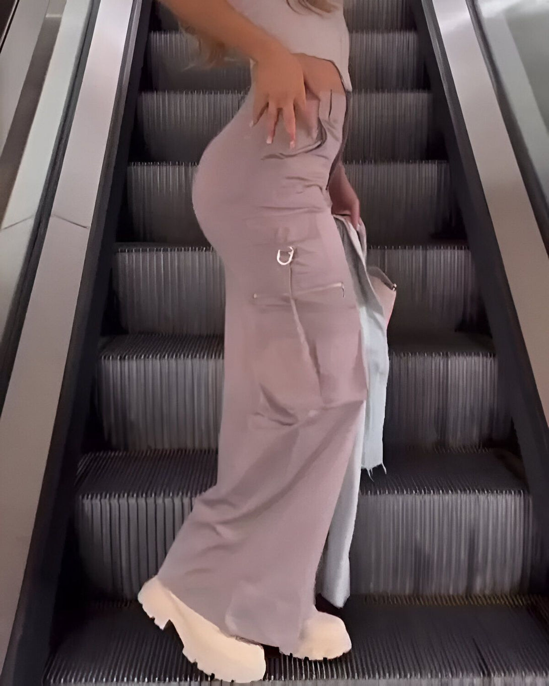 Une femme monte un escalator. Elle porte un pantalon beige et un haut court beige. Elle a une main sur sa hanche et l'autre tient un sac à main.