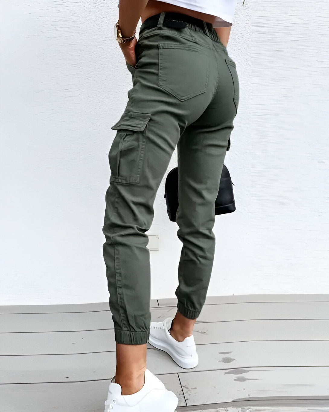 Une femme porte un pantalon cargo vert olive avec des poches cargo sur les côtés. Elle porte également un t-shirt blanc et des baskets blanches.